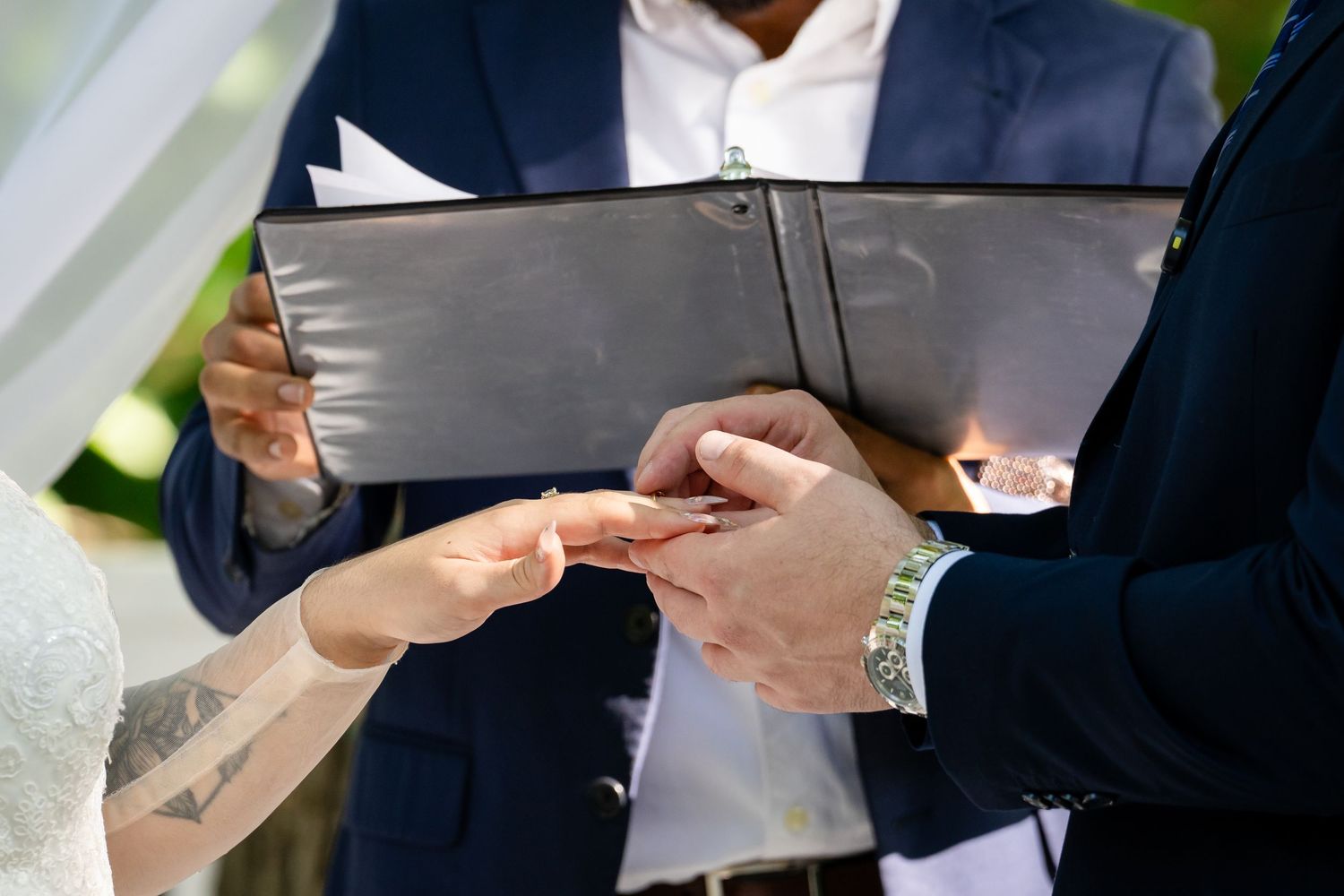 Groom placing a wedding ring on bride's finger during outdoor ceremony, officiant holding black binder in background.