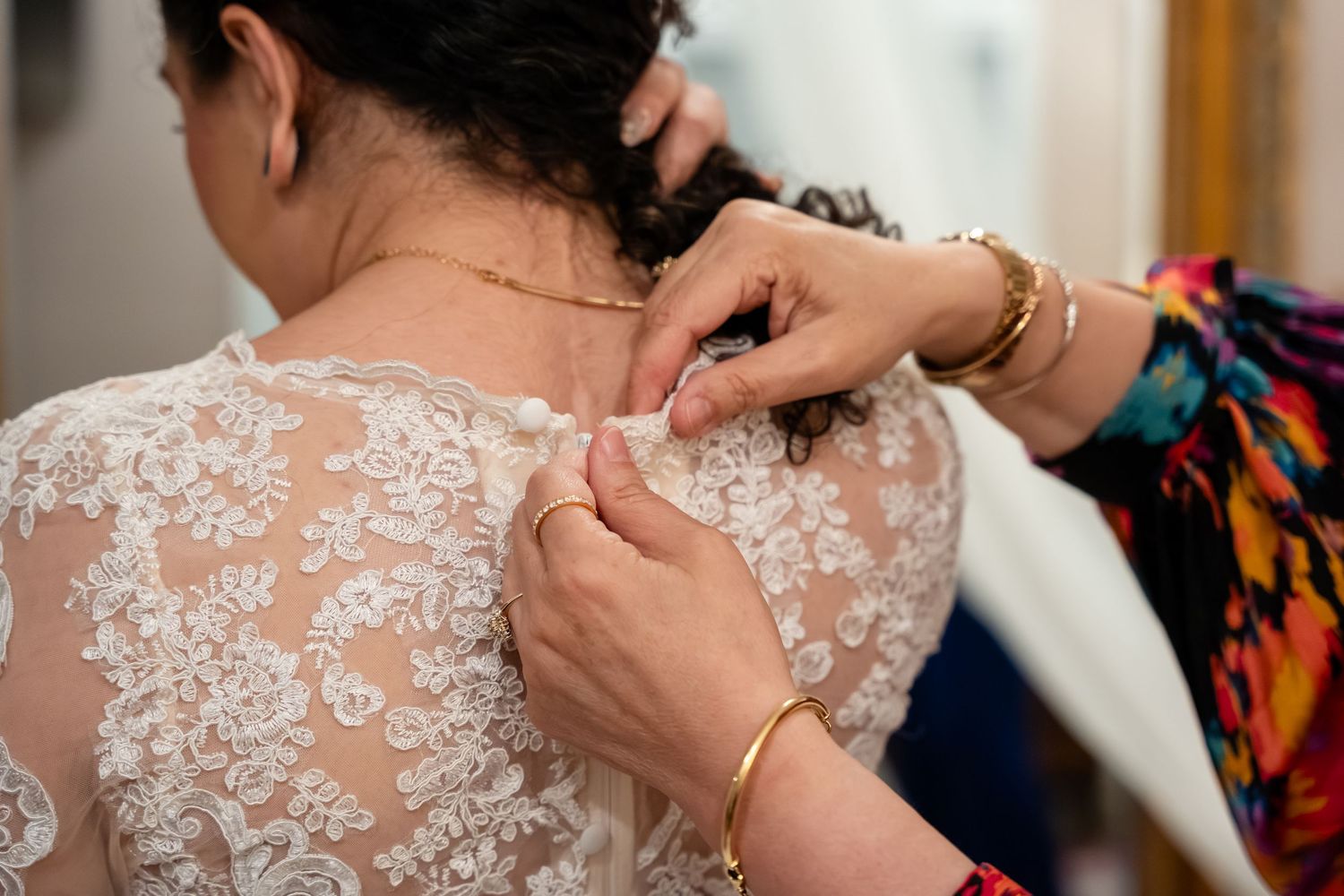 A bride in a lace wedding dress getting her back buttons fastened by a helper wearing a colorful floral outfit.