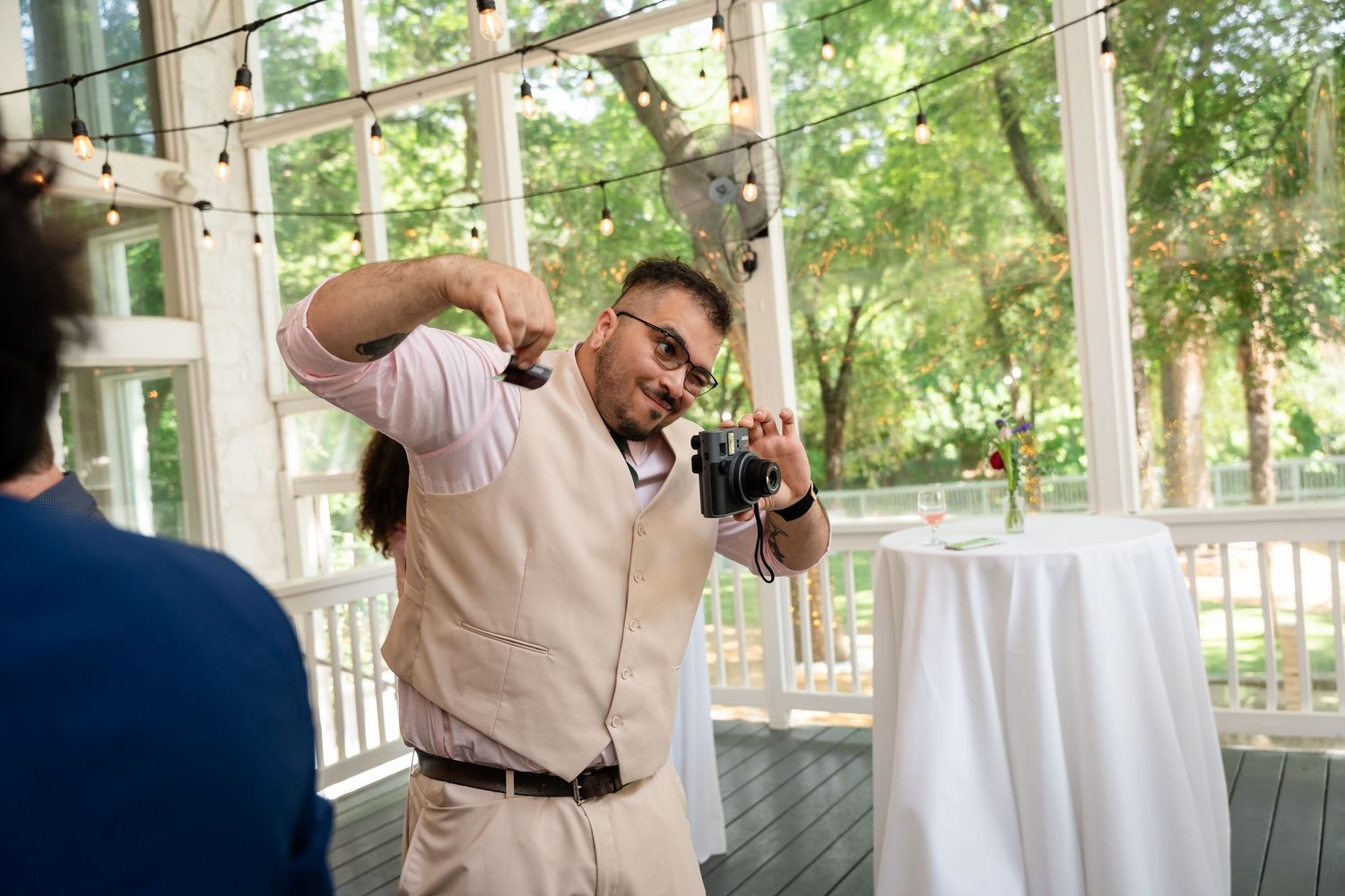 Photographer capturing moments at an outdoor wedding reception on a decorated porch with string lights and white cocktail tables.