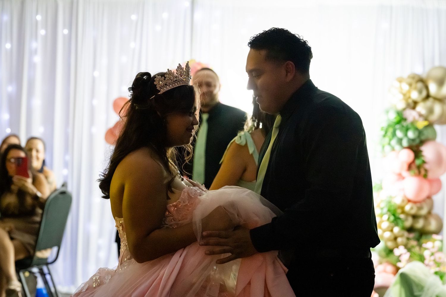 A young woman in a pink quinceañera gown and tiara shares a dance with a man in a black suit at her celebration.