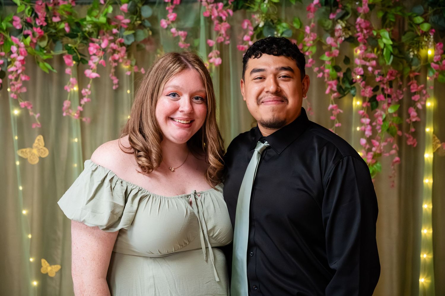 Couple posing together in front of floral backdrop with pink flowers and butterfly decorations at formal event.