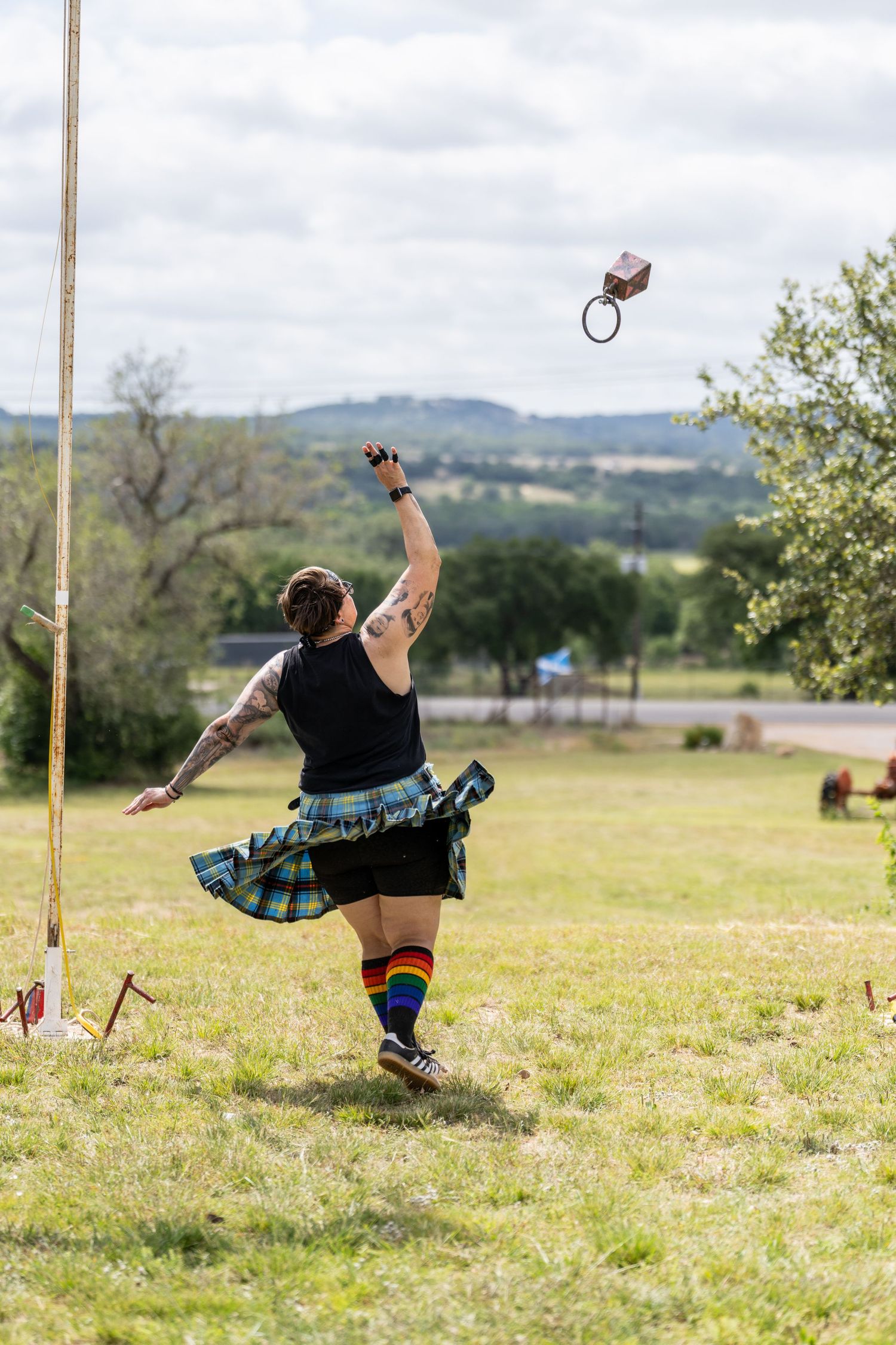 Athlete in tartan kilt and rainbow socks throws weight over bar at Scottish Highland Games outdoor competition.