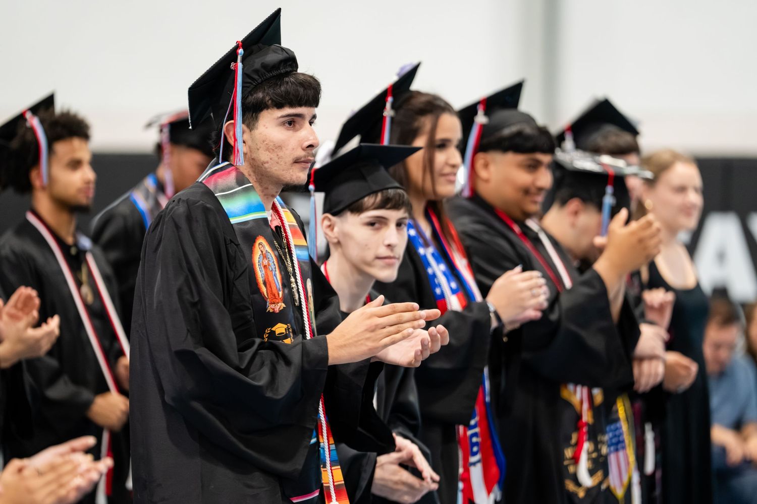 Graduates in black caps and gowns clapping at a commencement ceremony, wearing colorful honor stoles.
