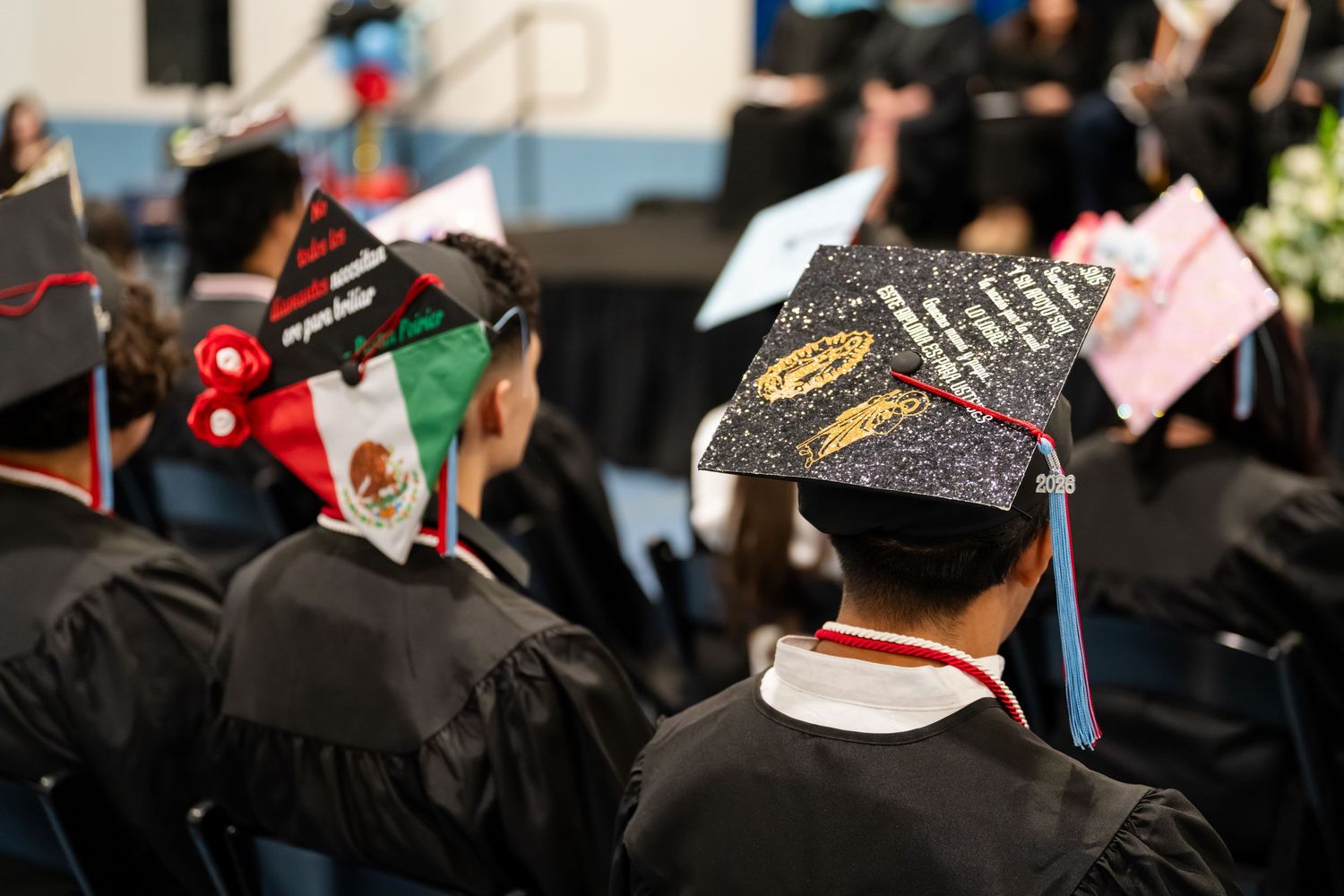 Graduates wearing decorated mortarboard caps at a graduation ceremony, including one with a Mexican flag design.