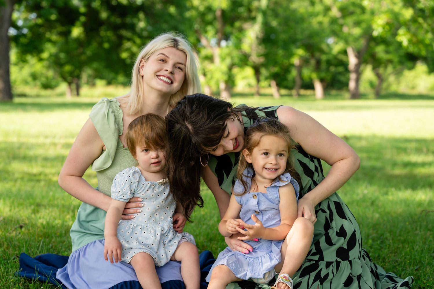 Two women and two little girls enjoy a sunny day together sitting on grass in a lush green park.