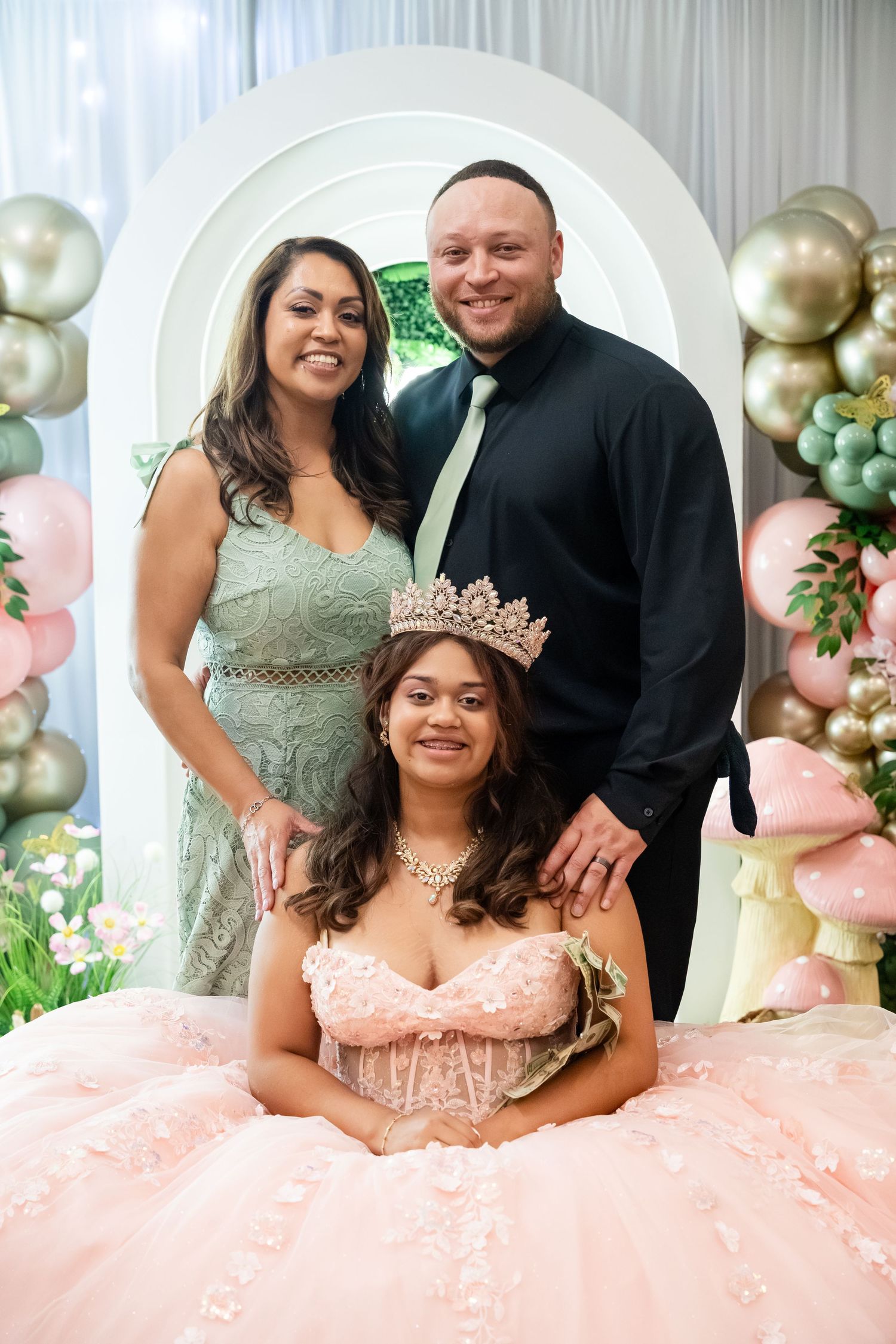 Family posing at a quinceañera celebration, girl in pink gown and crown seated with parents behind her.