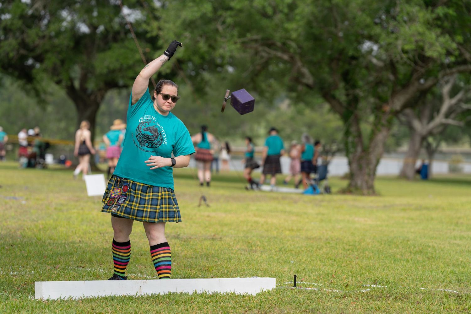 Person in tartan kilt and teal shirt throwing hammer at Highland Games competition in a grassy park.