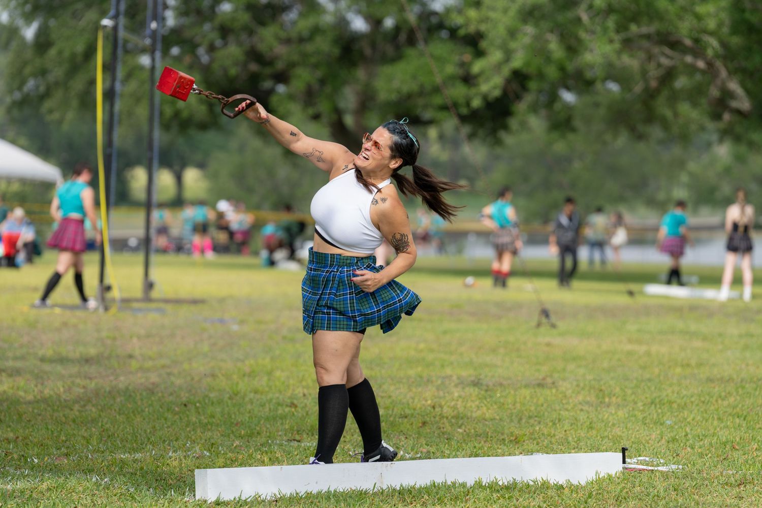 Woman in plaid skirt throwing a shot put at a Highland Games athletic competition outdoors.