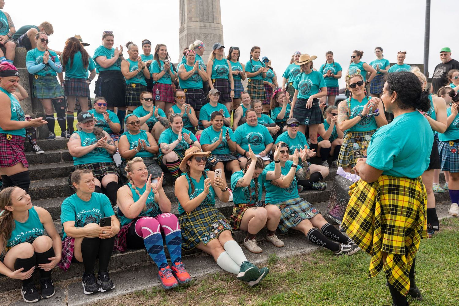 Large group in teal shirts and kilts clapping outdoors near a monument at a Scottish Highland gathering event.