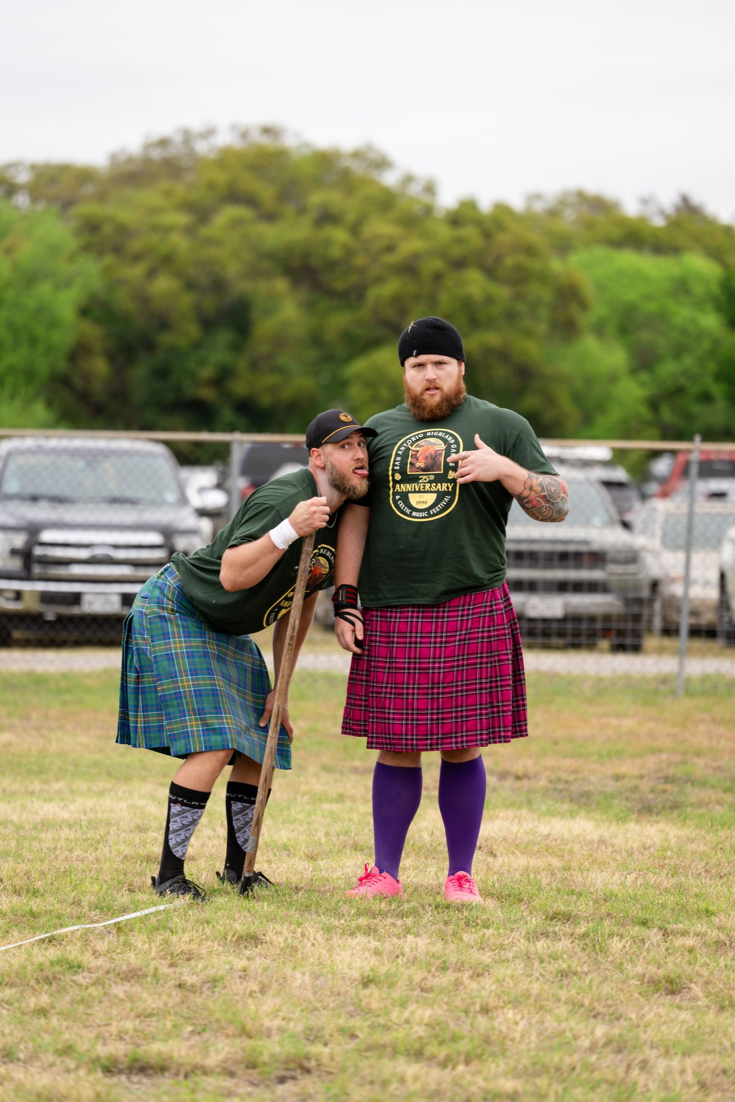 Two men in traditional Scottish kilts and green shirts pose together at a Highland Games event outdoors.