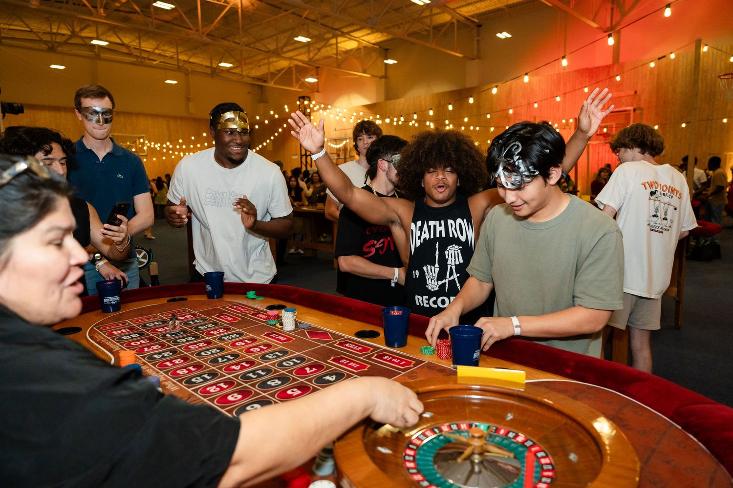 A group of players gathered around a roulette table at a casino-themed event with string lights overhead.