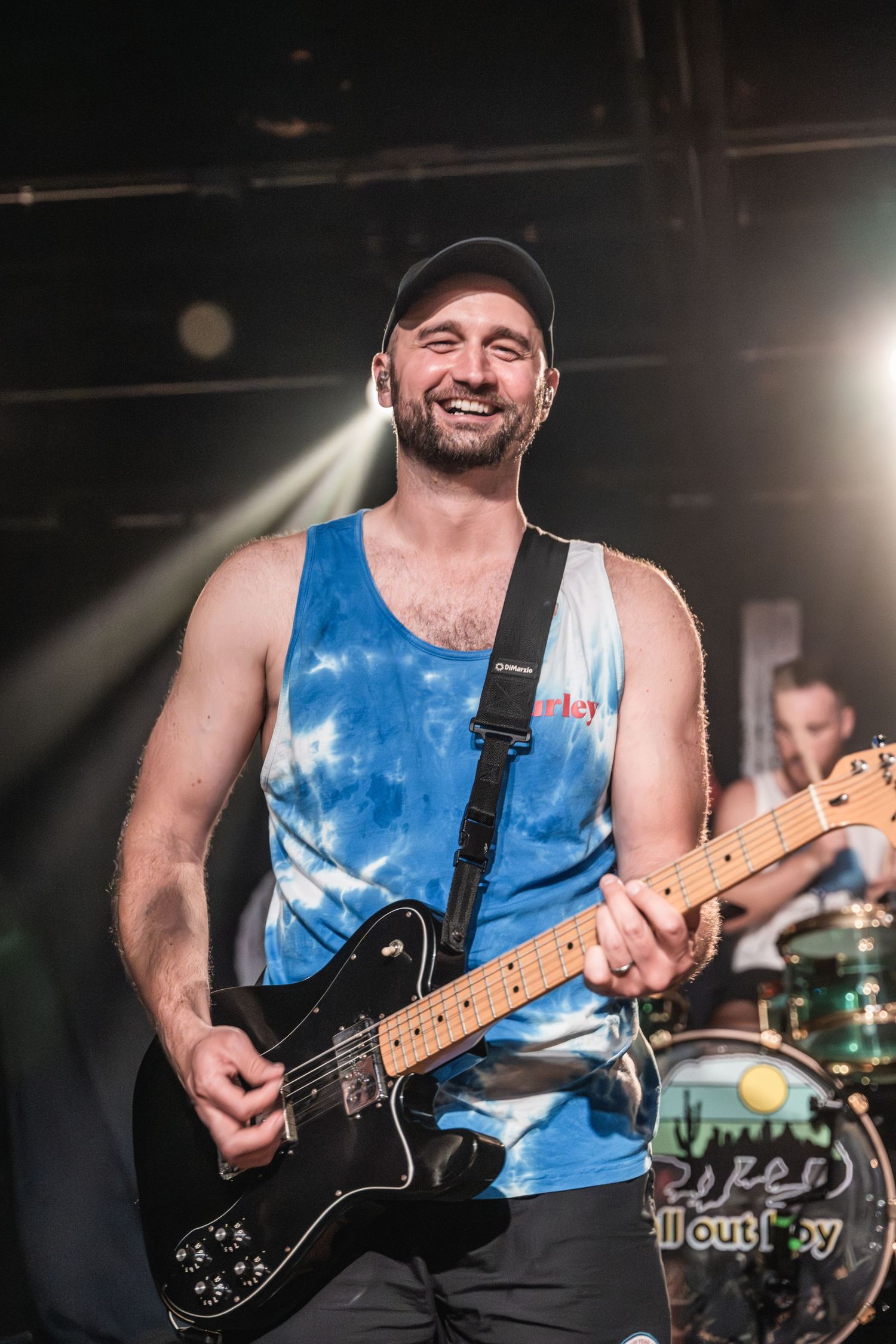 Guitarist in blue tie-dye tank top performs on stage with spotlights.