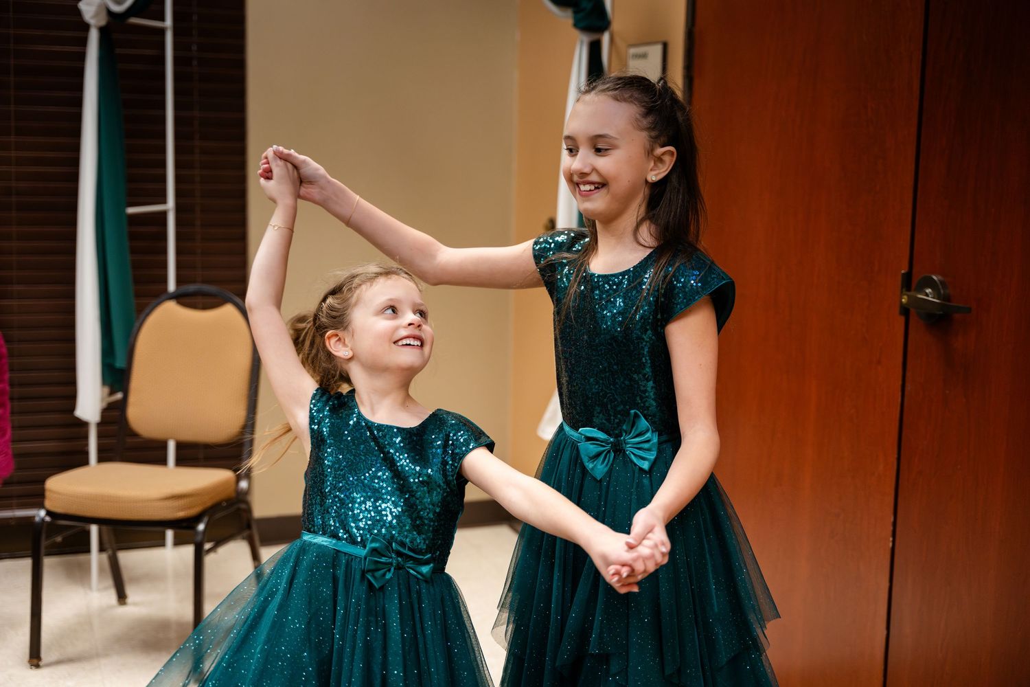 Two dancers in matching teal sequined dresses share a joyful moment holding hands before a performance.