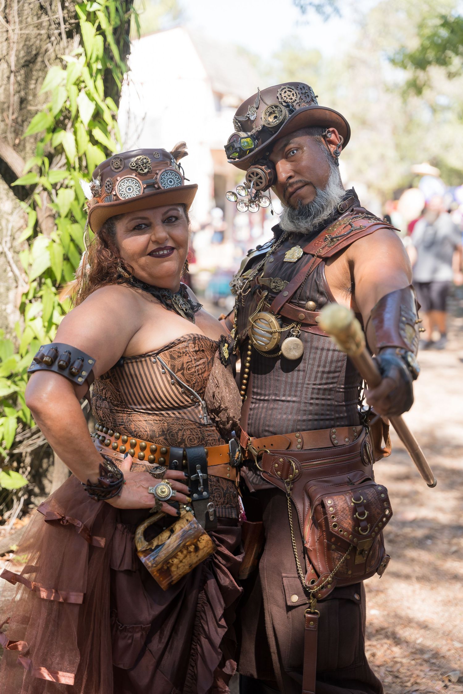 A couple poses in steampunk-inspired costumes featuring leather and metal accessories at an outdoor festival.