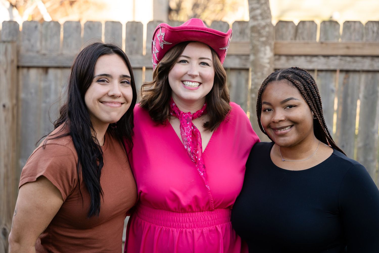 Three smiling friends standing together outdoors against a wooden fence, with one wearing a bright pink dress and hat.