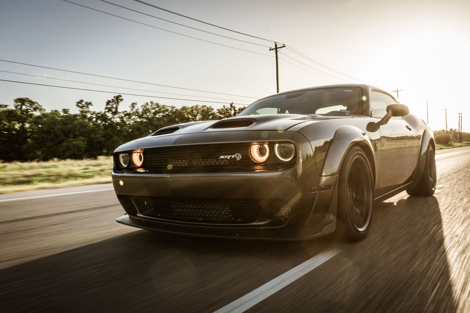 Black Dodge Challenger with custom modifications speeds down a sunlit road.