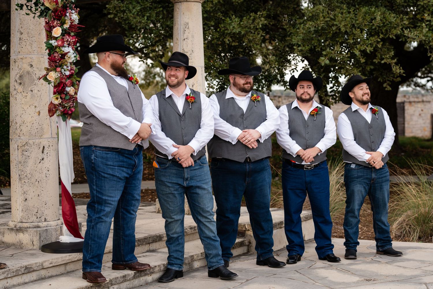 Five groomsmen in western-style wedding attire stand in line at an outdoor venue with stone columns.