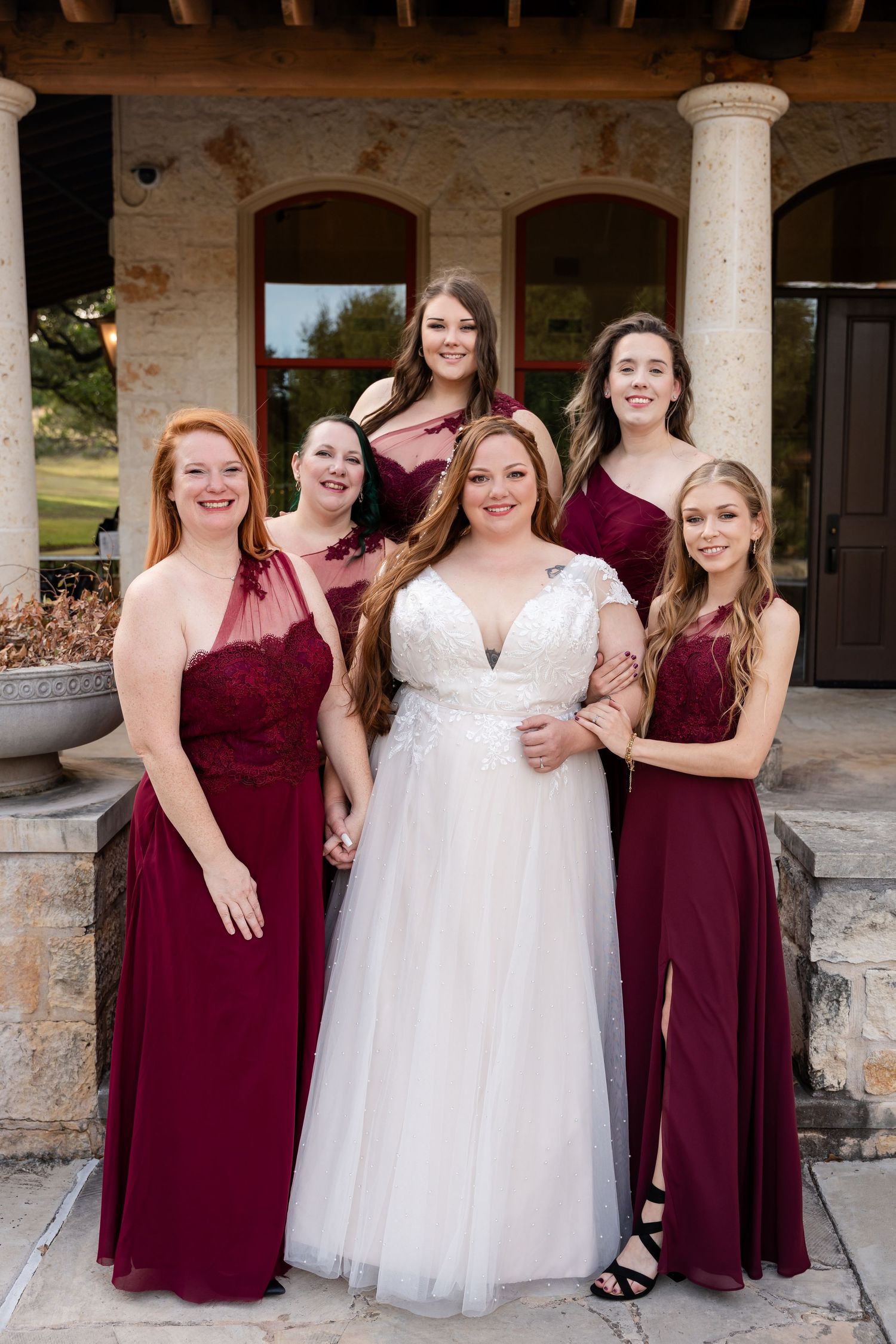 Bridal party in burgundy dresses posing on stone porch with columns.