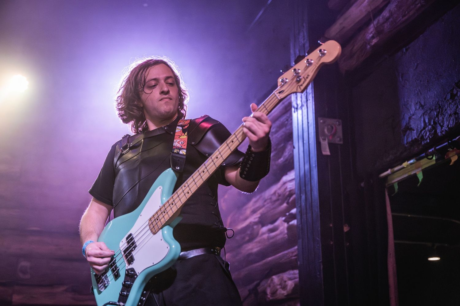 Bass guitarist plays turquoise instrument under purple stage lighting during live performance.