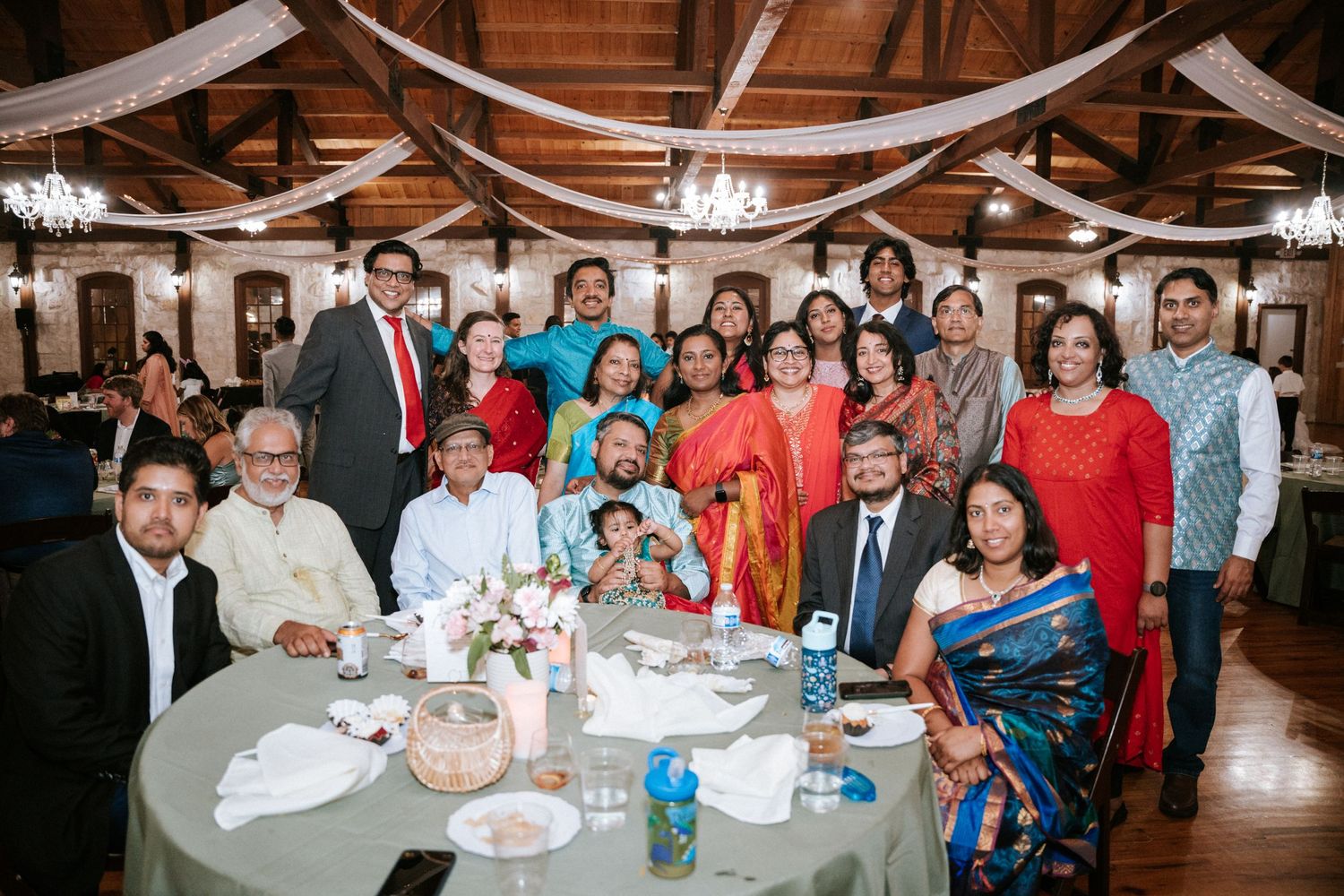 A large family group poses together at a wedding reception table decorated with centerpieces.