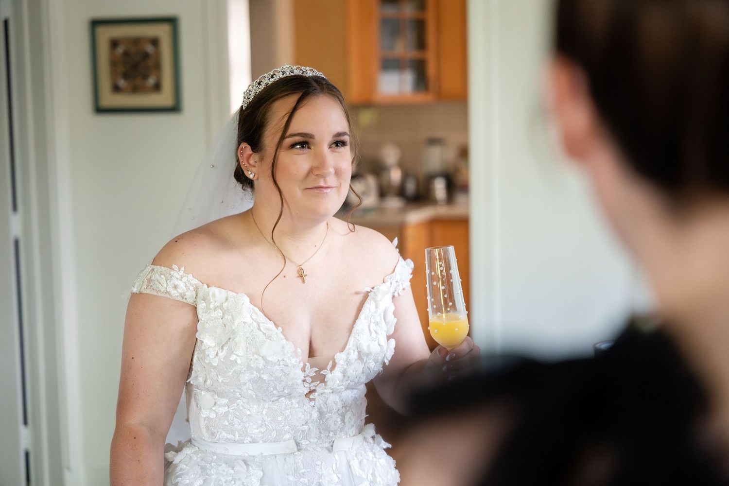 A person in an elegant white lace wedding dress stands near a window in soft natural light.