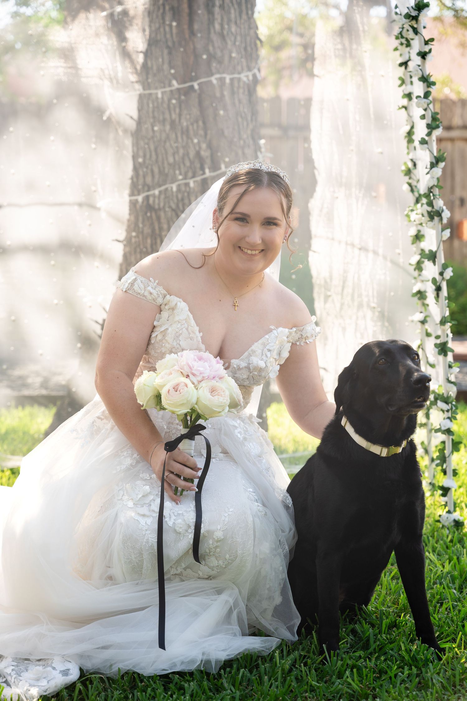 A bride in lace gown poses with black Labrador dog under string lights in garden setting.