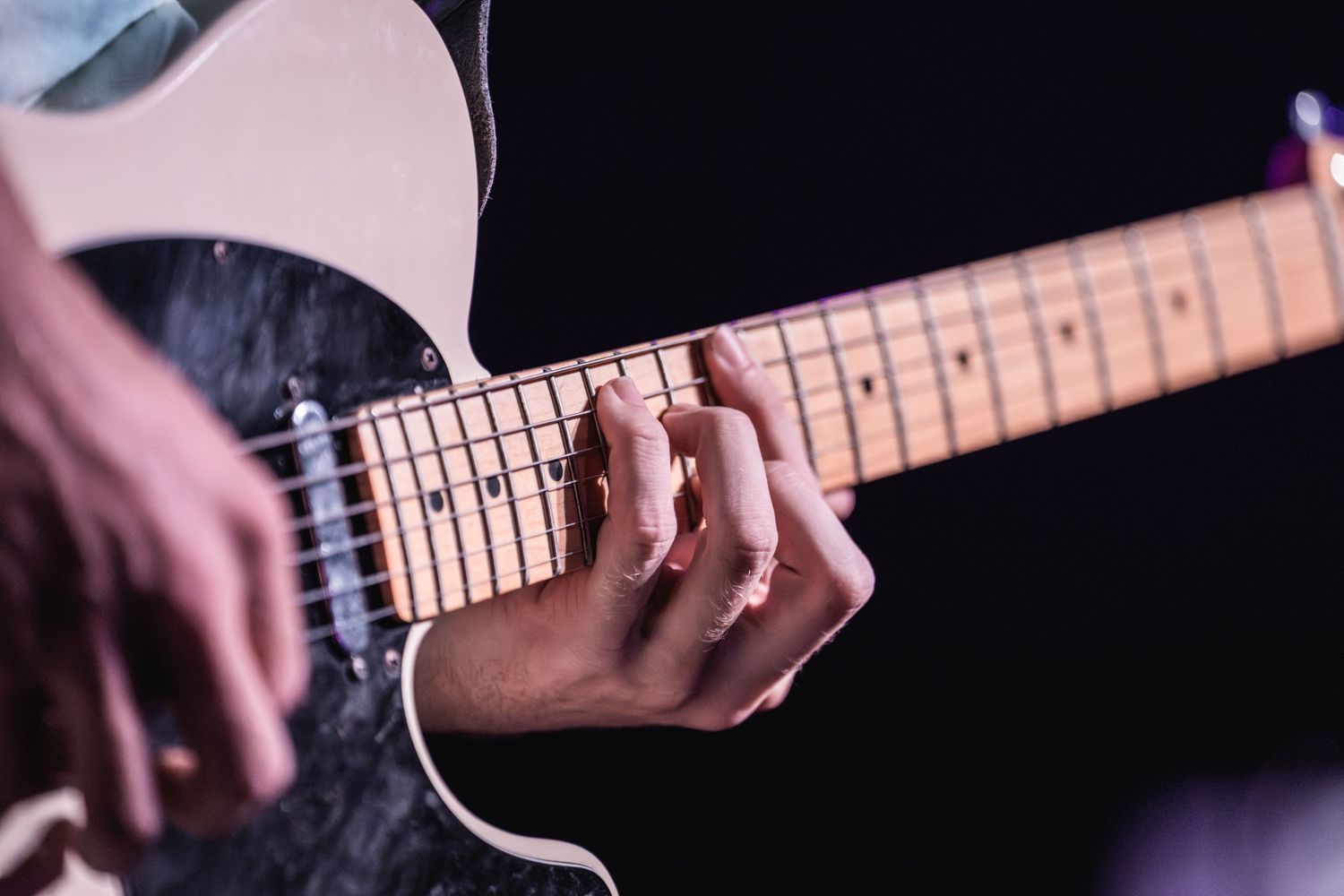 Close-up shot of hands playing electric guitar with maple fretboard against dark background.