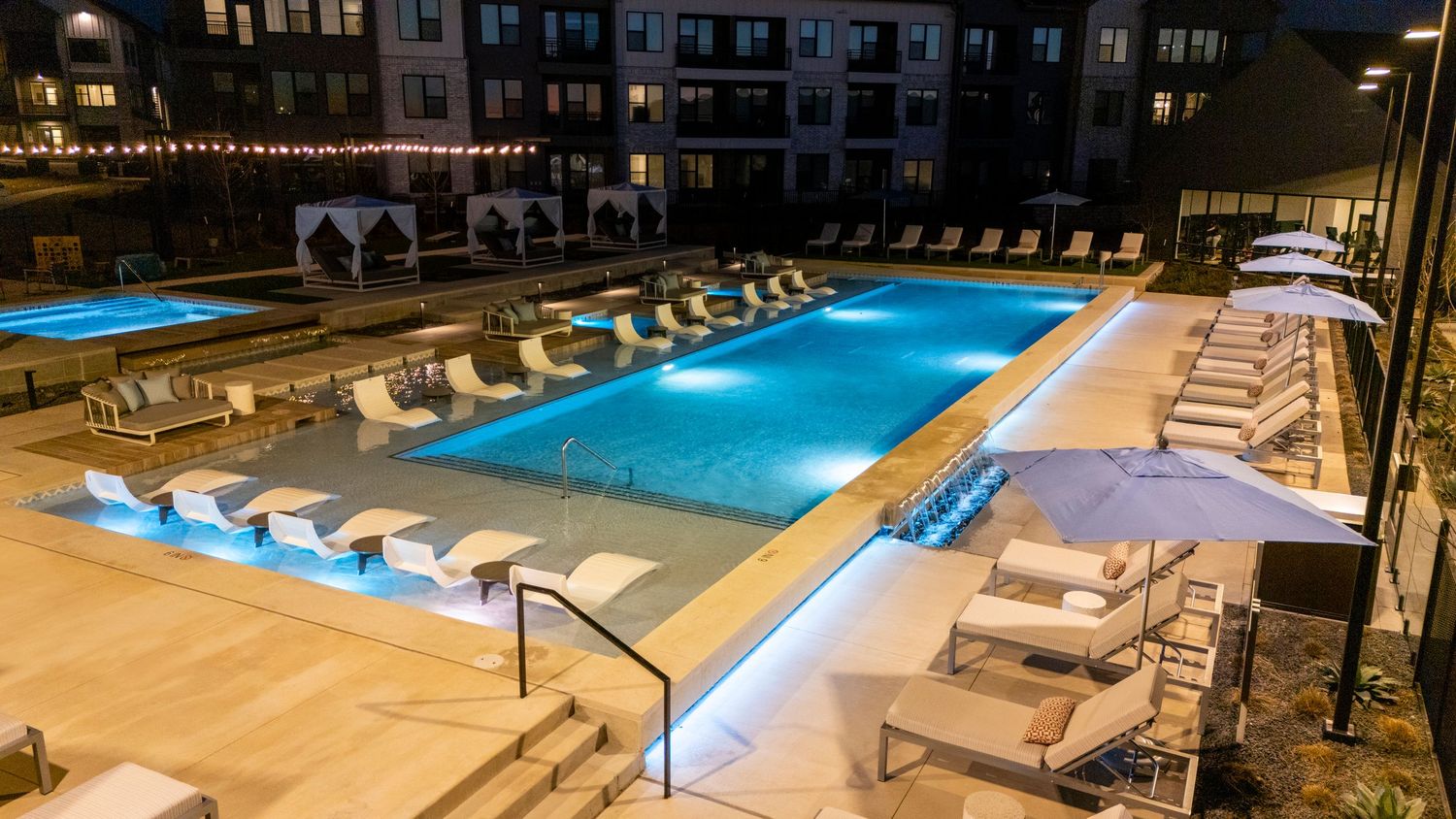 Illuminated luxury apartment complex pool deck with lounge chairs and modern lighting at dusk.