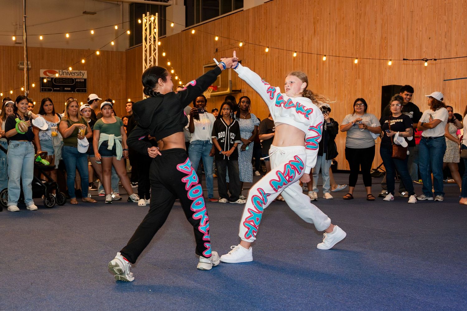 Two dancers perform a dynamic dance routine in front of a crowd at night with string lights in the background.