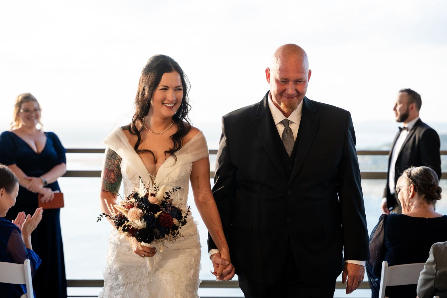 Newlyweds exit ceremony holding hands with guests looking on at waterfront venue.