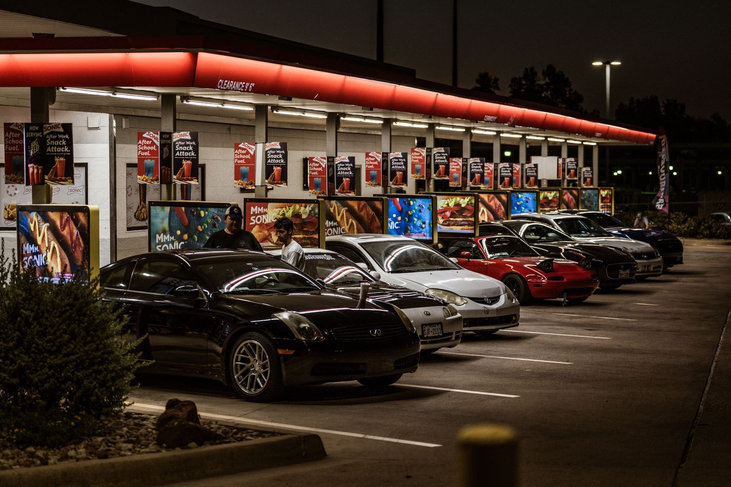 Line of luxury sports cars parked under illuminated red canopy at night.