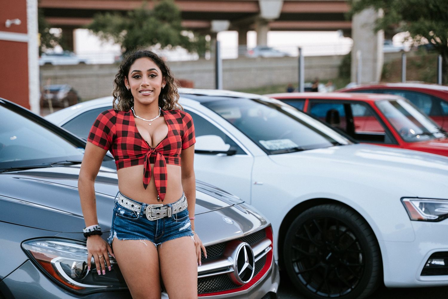 Red plaid tied shirt and denim shorts outfit showcased with luxury cars in background.