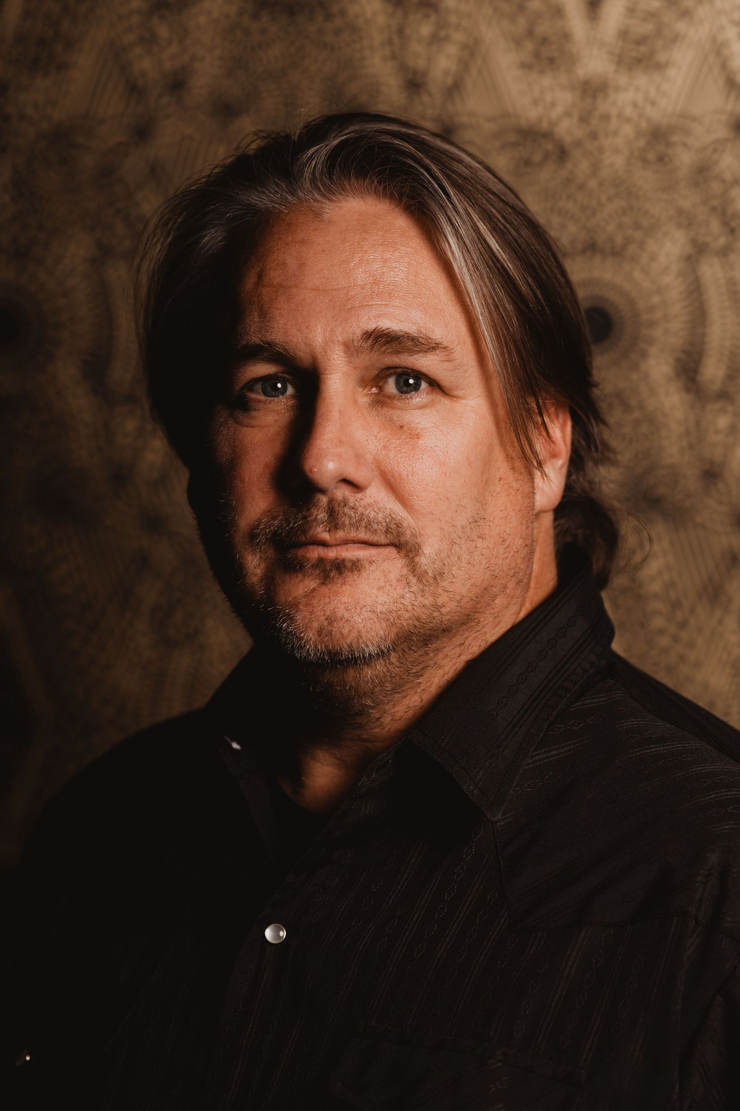 Artistic headshot of a man with long hair in black shirt against pattern background.