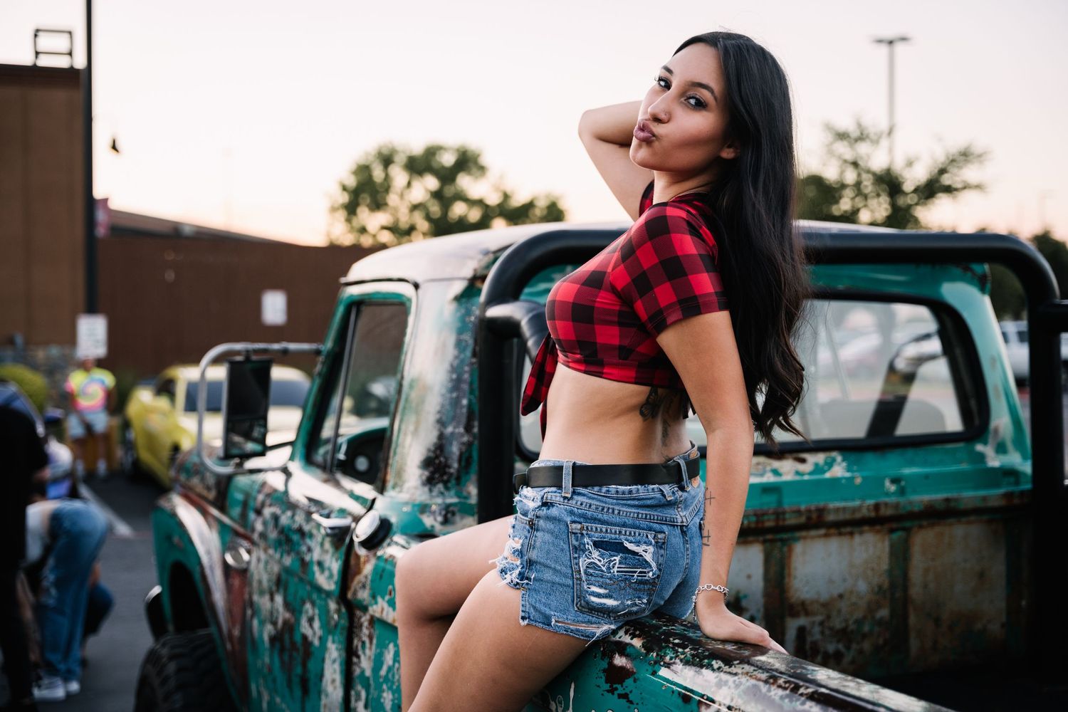 A model poses on a vintage truck wearing a red plaid crop top and denim shorts at a car show.