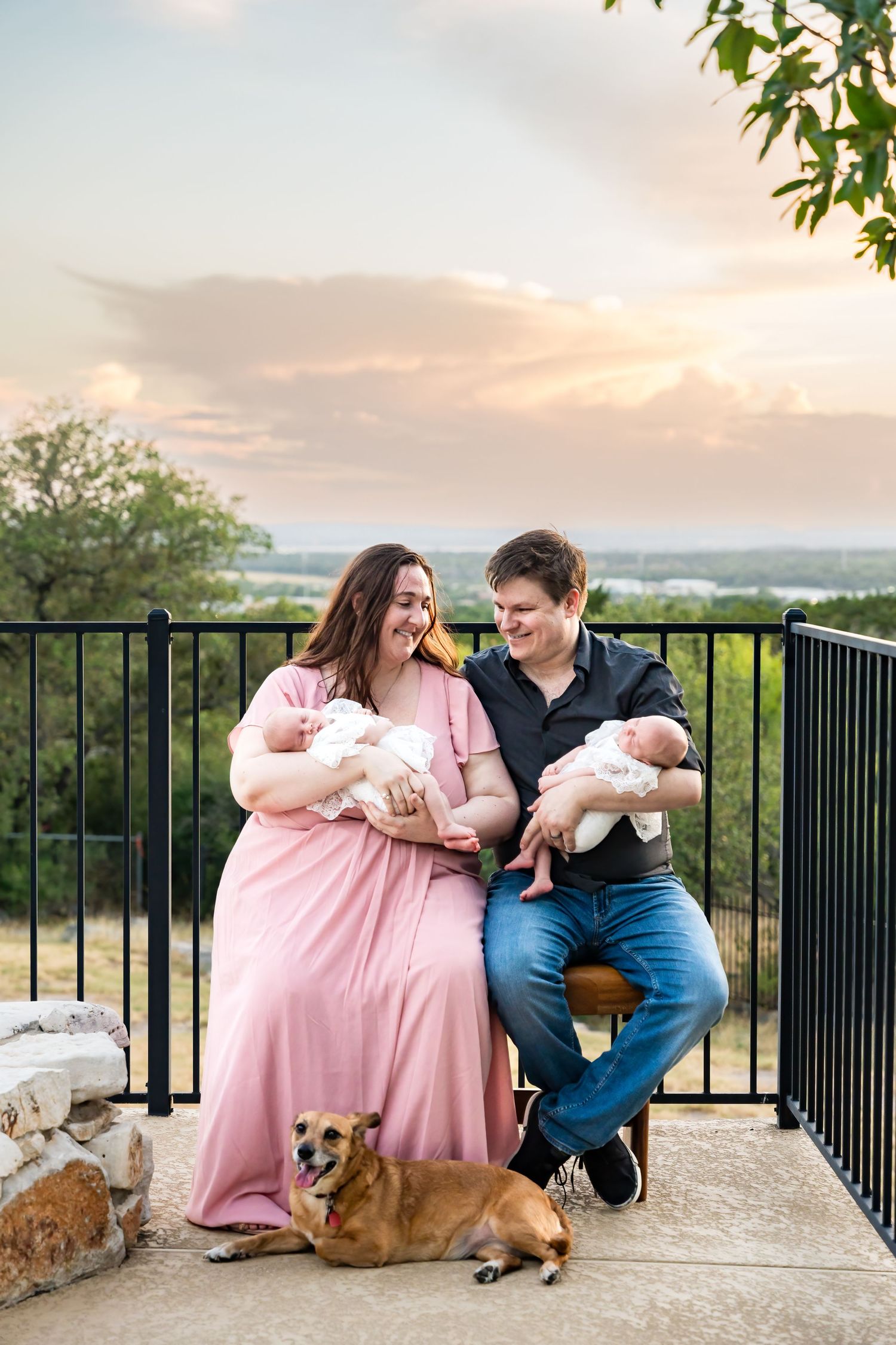 Couple sitting with newborn twins and dog on outdoor patio during sunset.