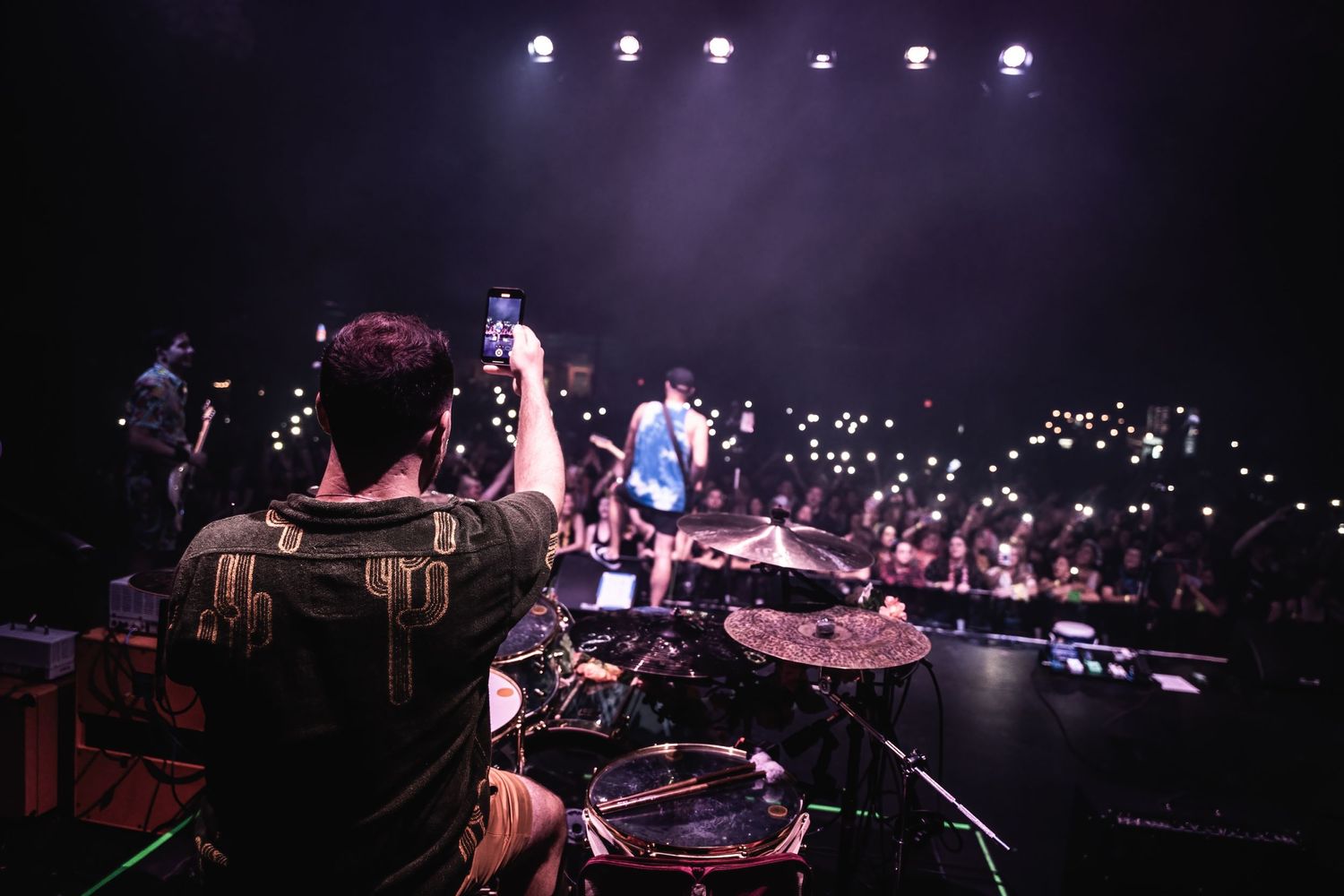 Drummer performs on stage at concert with dramatic lighting and crowd visible in background.