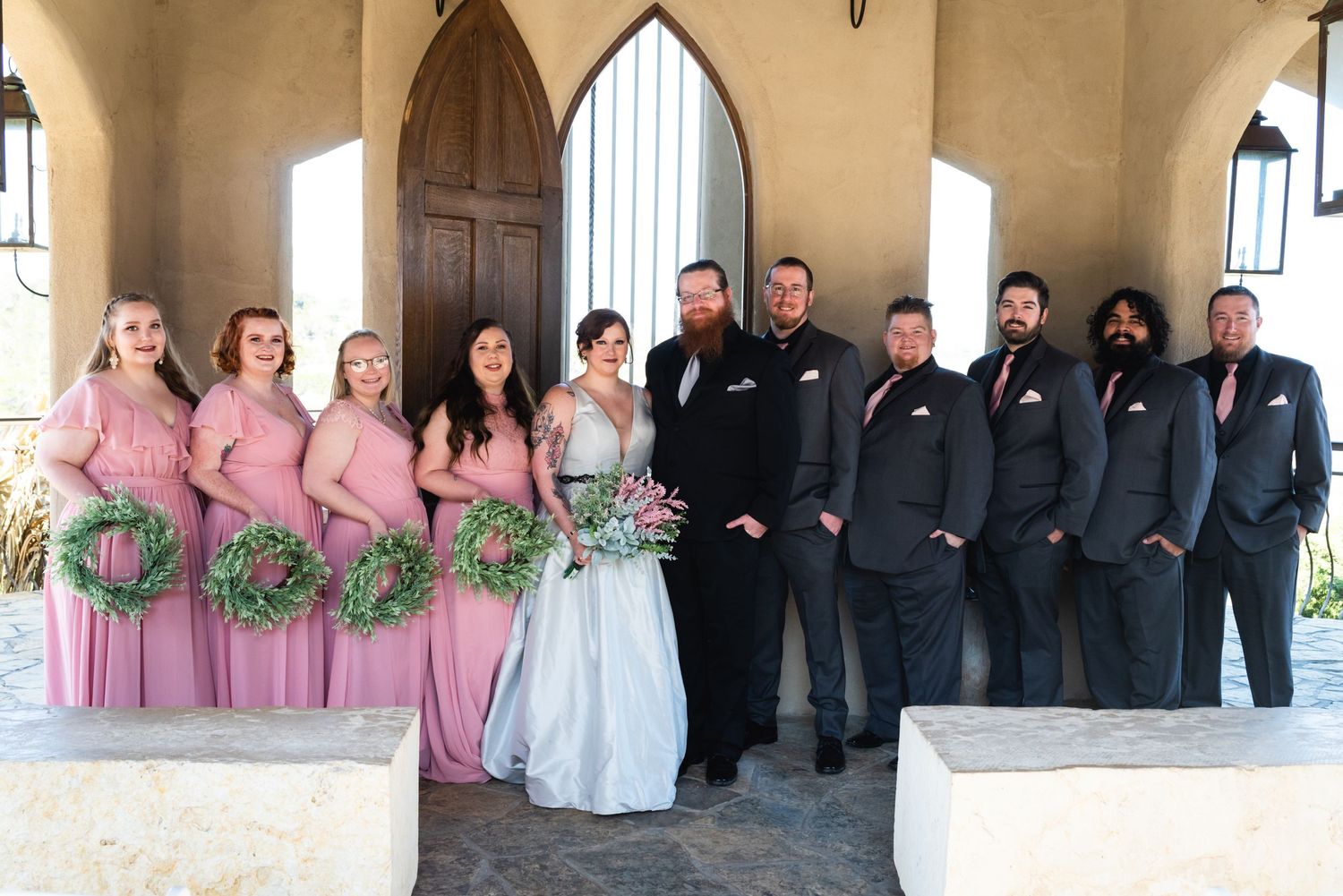 Wedding party in pink dresses and black suits pose together under an archway at a church.