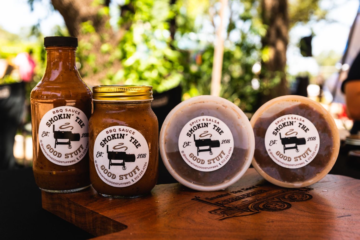 Branded barbecue sauce and rub products displayed on a wooden surface in sunlight.