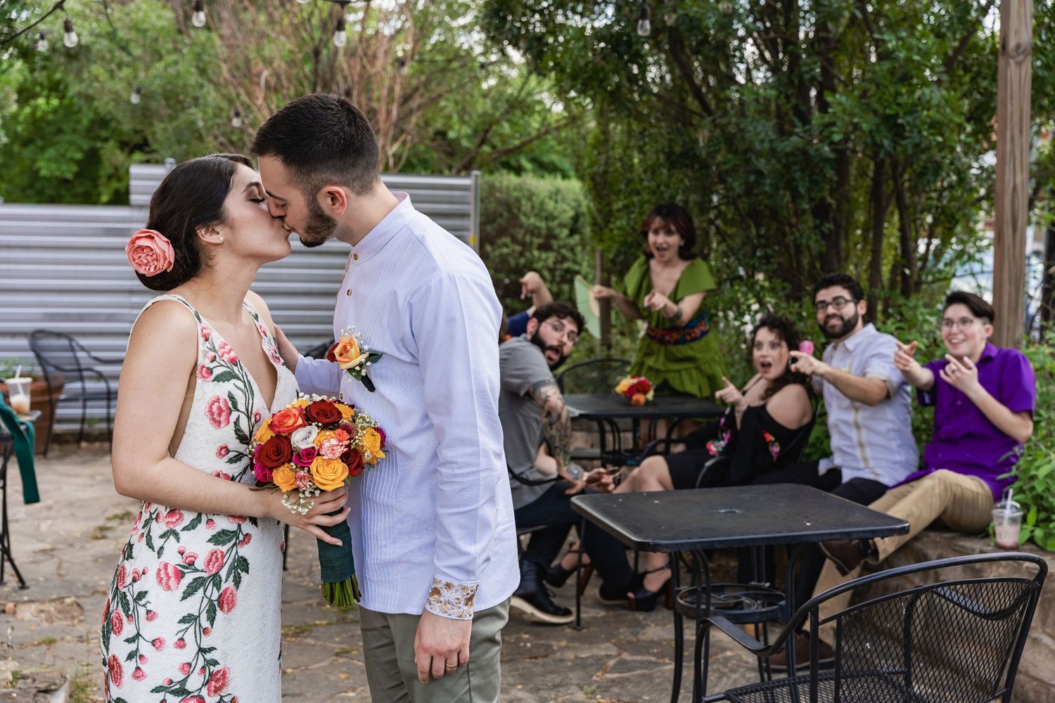 A couple shares a romantic moment in an outdoor courtyard while guests look on from nearby tables.
