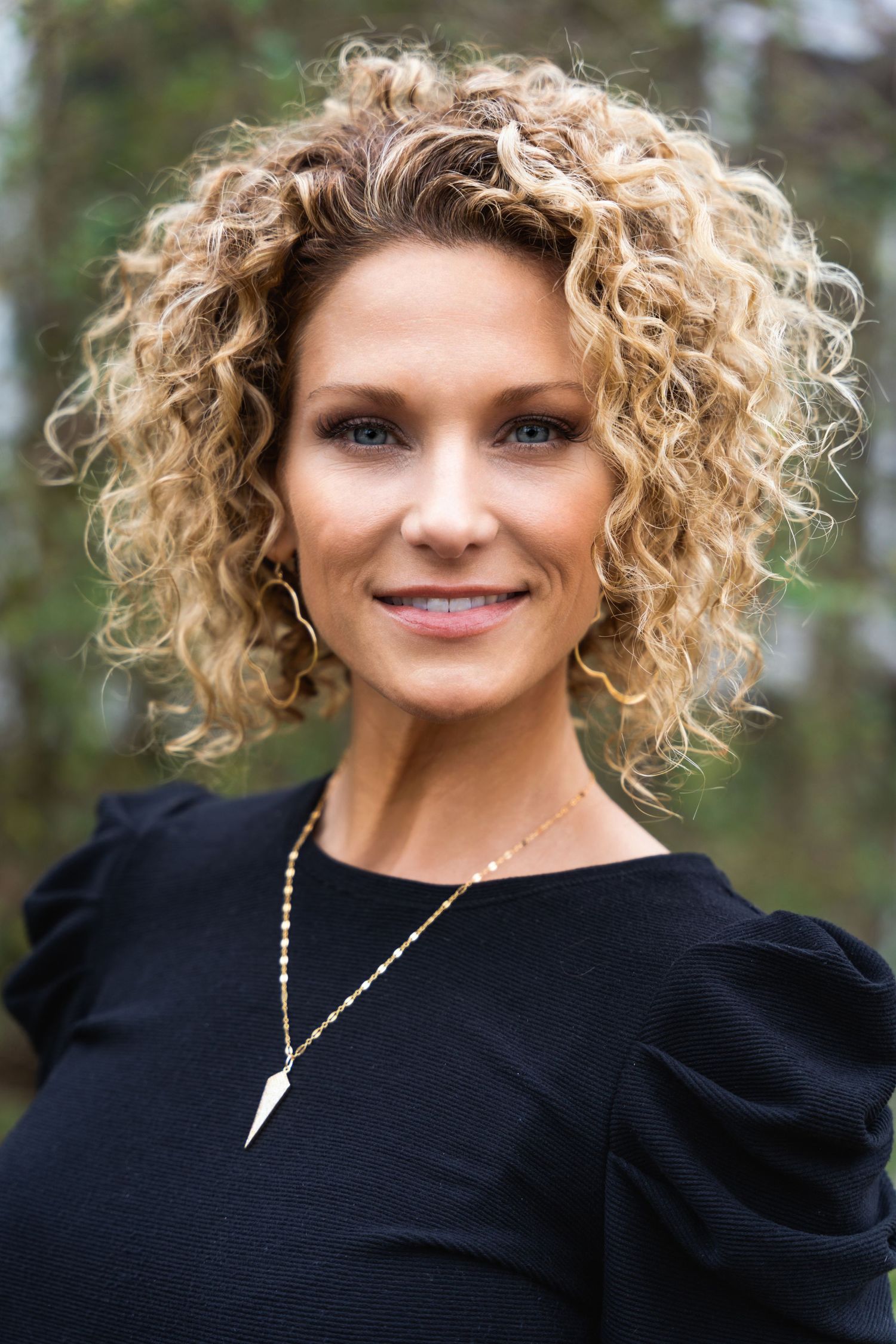 Professional headshot of person in black top with curly blonde hair and pendant necklace against natural background.