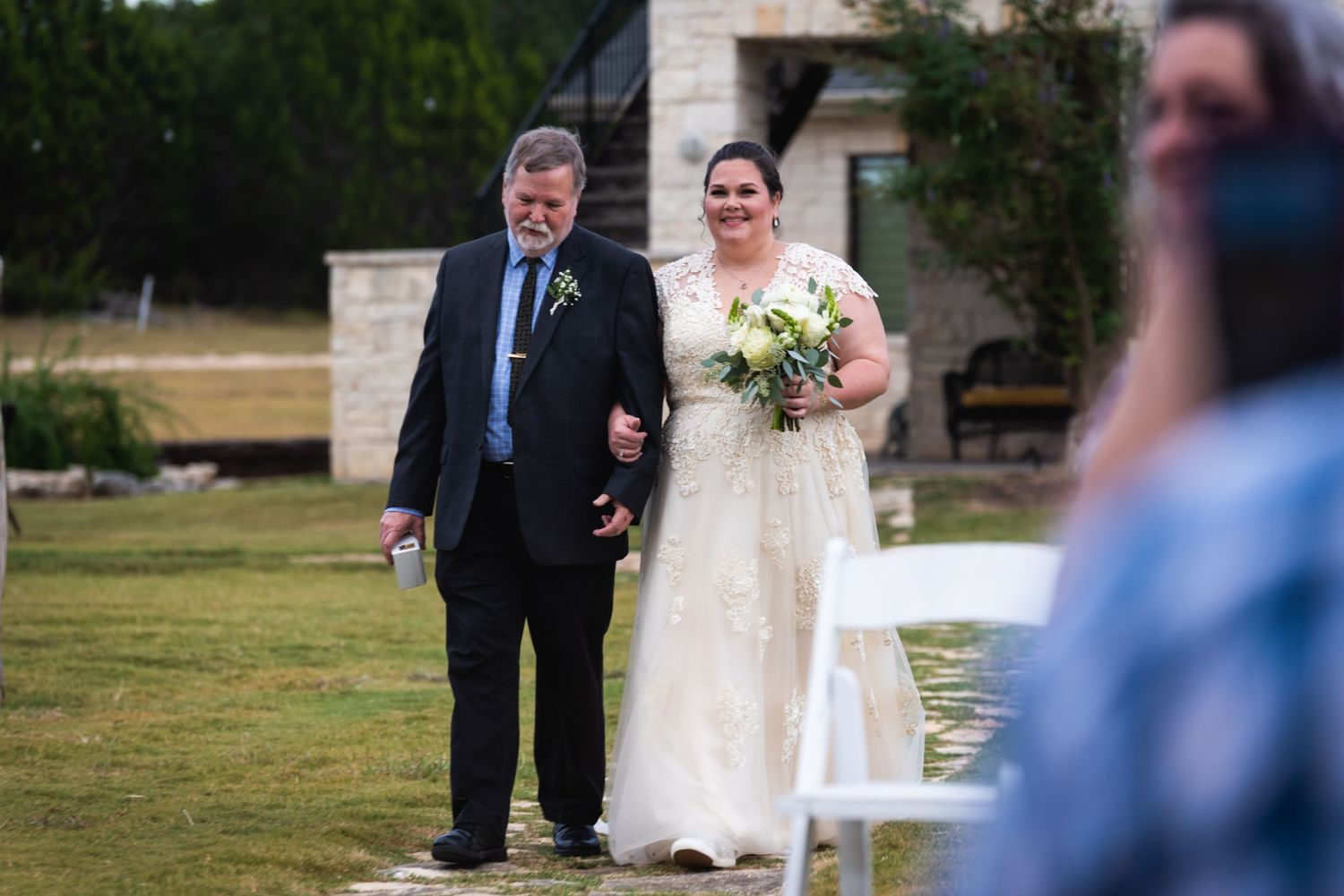 A bride in a white wedding dress walks down an outdoor aisle carrying a bouquet.