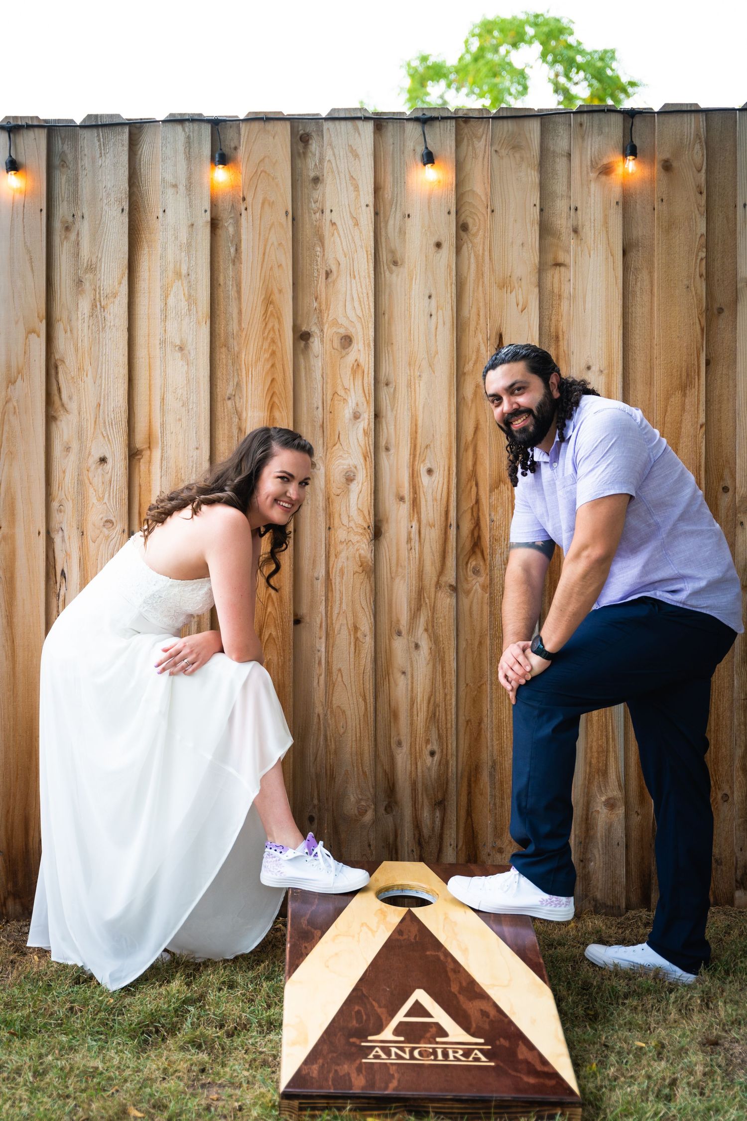 Wedding couple playing cornhole game against wooden fence with string lights.