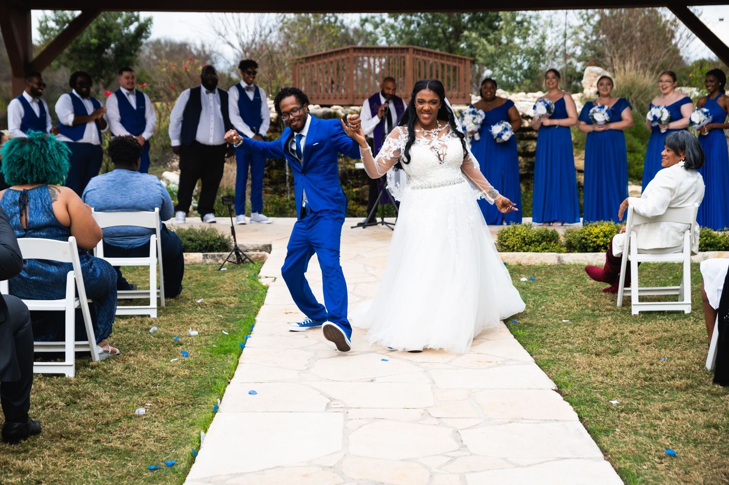 Wedding party members dance joyfully down an outdoor aisle in blue attire and white dress at a garden ceremony.