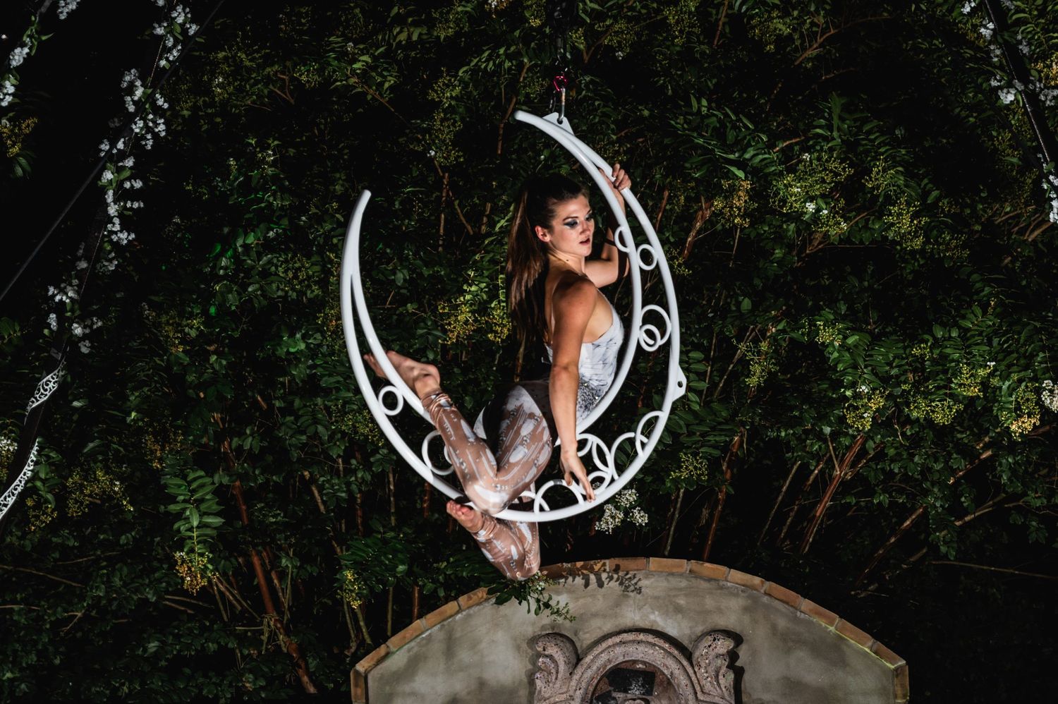 Performer poses gracefully on a decorative crescent moon prop against dark foliage.