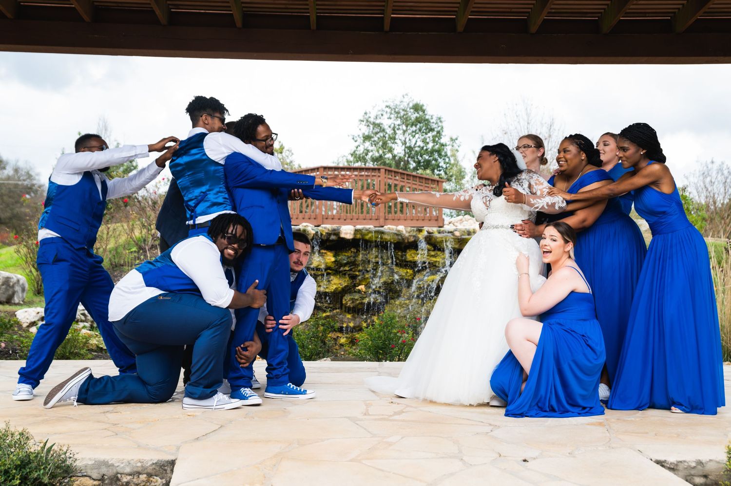 Wedding party poses in playful tug-of-war with bride and bridesmaids in royal blue against groomsmen on outdoor patio.