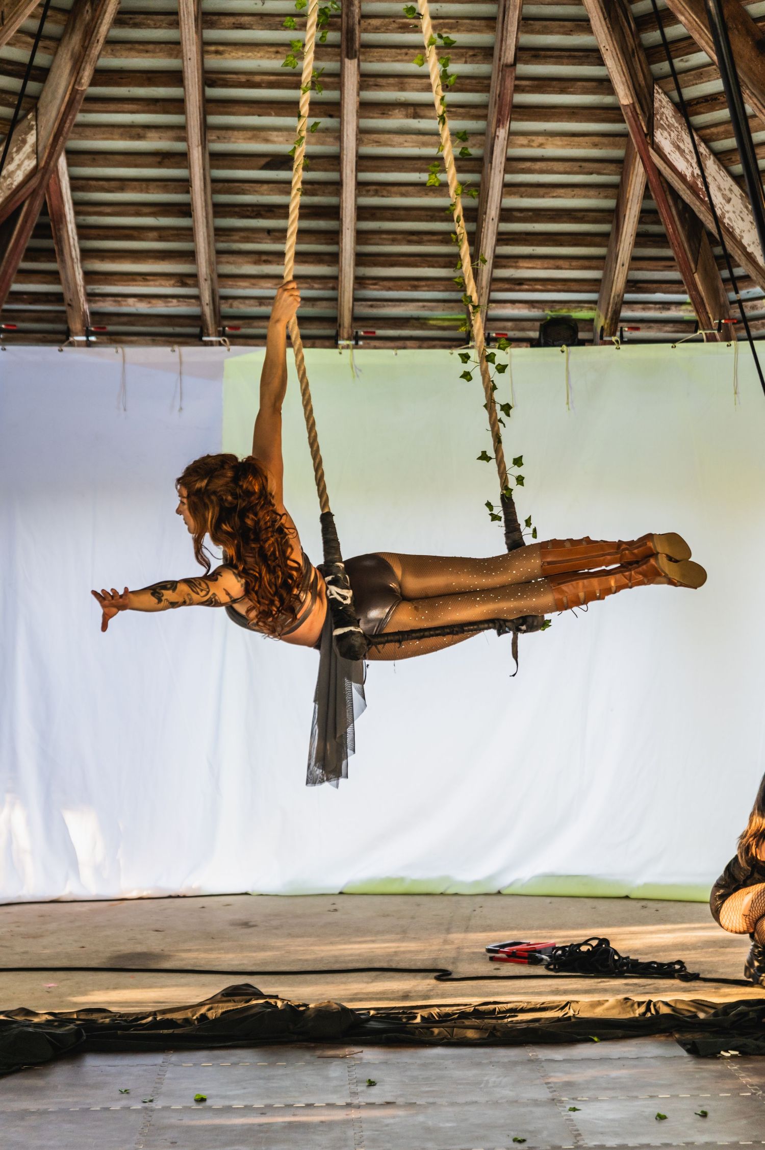 Aerial performer practices on suspended equipment in a rustic indoor space with wooden beams.