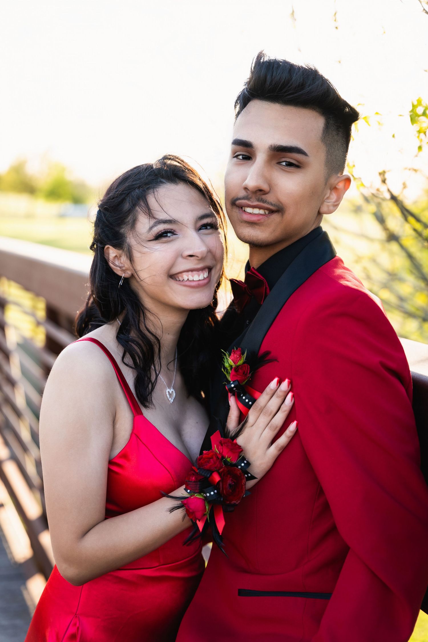 Young couple in matching red formal attire posing together with floral corsage on an outdoor bridge.