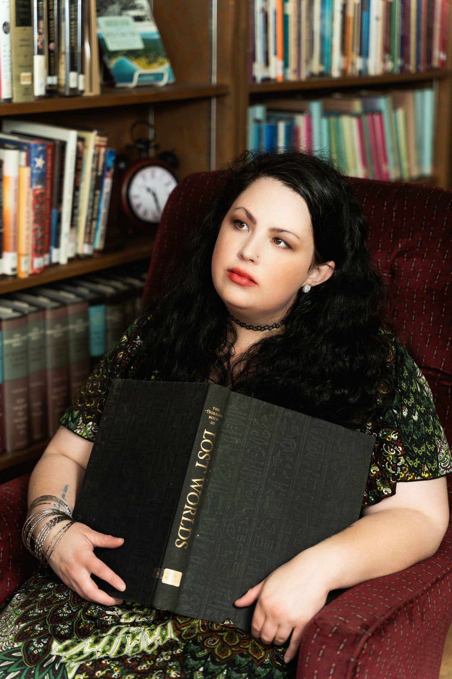 Portrait in library setting holding large book while seated in vintage armchair.