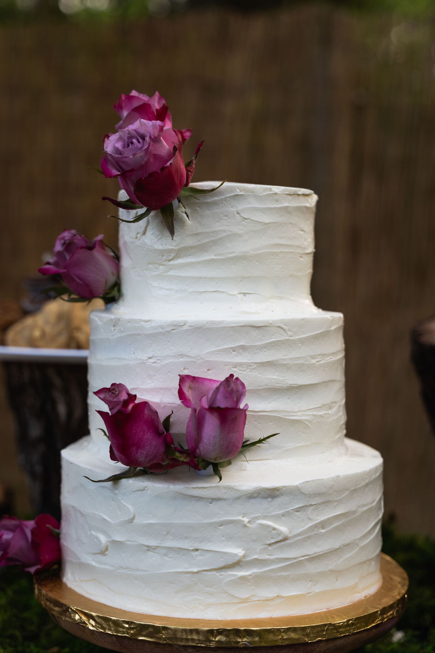 Three-tiered white wedding cake decorated with purple roses on a gold stand.