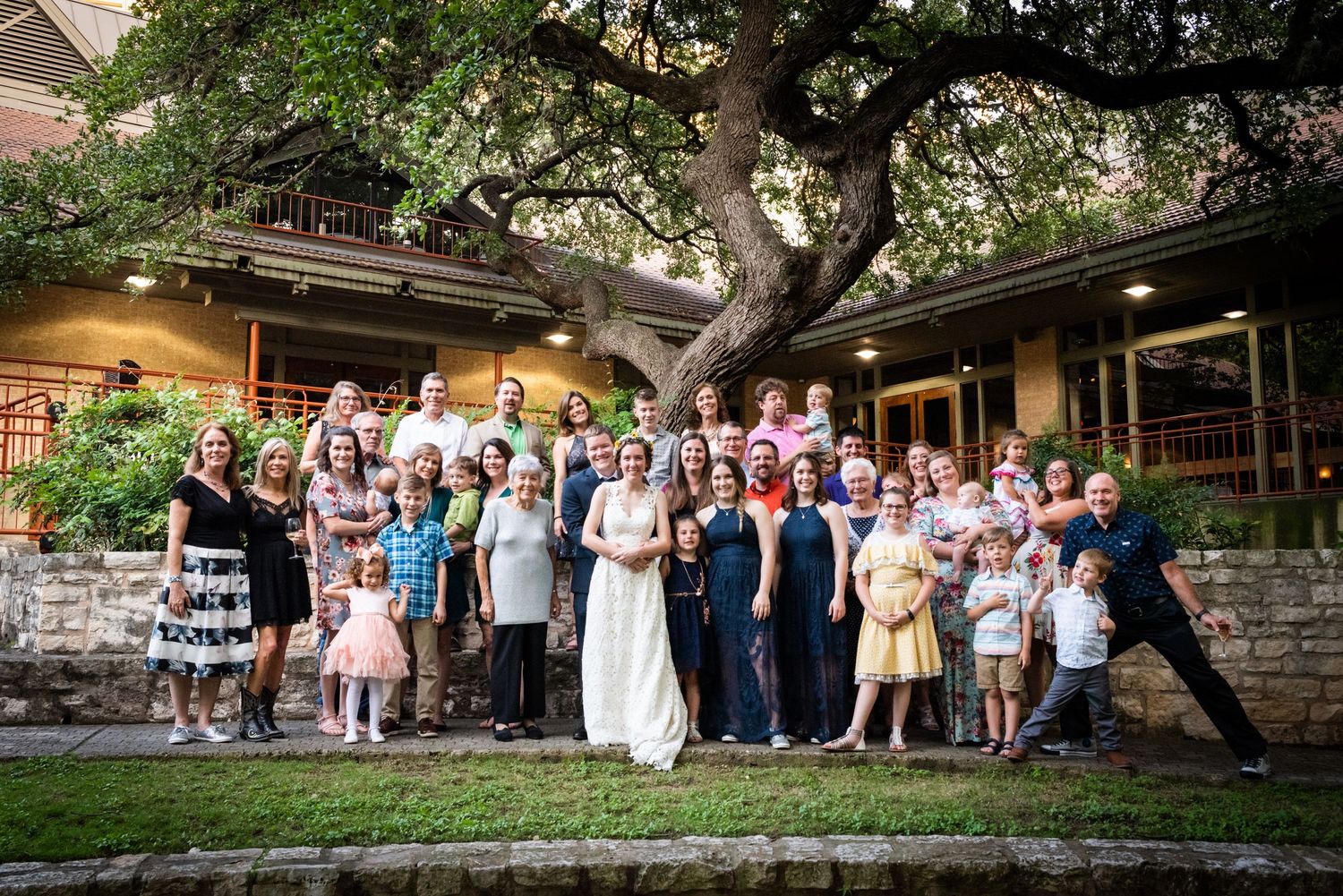 Large wedding party group photo taken outdoors under a sprawling tree.
